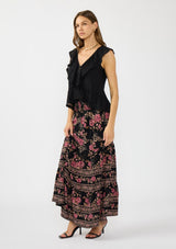 [Color: Black/Dusty Rose] A side facing image of a brunette model wearing a black and pink maxi skirt with a smocked waist and a flowy, tiered silhouette. Styled with a black ruffled top. 