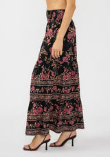 [Color: Black/Dusty Rose] A side facing image of a model wearing a black and pink maxi skirt with a smocked waist and a flowy, tiered silhouette. 