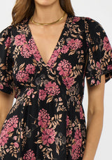 [Color: Black/Dusty Rose] A detail image of a brunette model wearing a black and pink floral print midi dress with short sleeves, a v neckline, and a handkerchief hemline. 