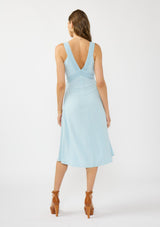 [Color: Blue] A back facing image of a brunette model wearing a light blue sleeveless midi dress with lace trim accents, a v neckline, and an empire waist. 