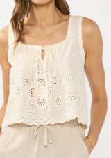 [Color: Cream/Natural] A detail image of a brunette model wearing a beige eyelet tank top with a square neckline, tie detail, and a scalloped trim. 