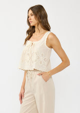 [Color: Cream/Natural] A side facing image of a brunette model wearing a beige eyelet tank top with a square neckline, tie detail, and a scalloped trim. 