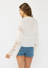 [Color: Off White] A back facing image of a brunette model wearing a white lace patchwork bohemian blouse with long bell sleeves and a v neckline. 