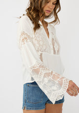 [Color: Off White] A side facing image of a brunette model wearing a white lace patchwork bohemian blouse with long bell sleeves and a v neckline. 