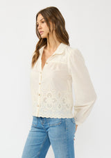 [Color: Cream/Natural] A side facing image of a brunette model wearing an off white embroidered shirt with a collared neck and button front. 