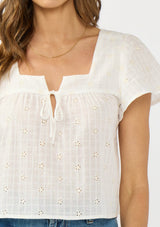 [Color: Natural/Cream] A detail image of a brunette model wearing a white embroidered crop top with short flutter sleeves and a square neckline with tie detail.