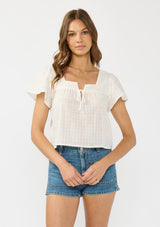 [Color: Natural/Cream] A front facing image of a brunette model wearing a white embroidered crop top with short flutter sleeves and a square neckline with tie detail.