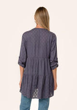 [Color: Purple Haze] A back facing image of a blonde model wearing a silky bohemian tunic top in a dusty purple diamond jacquard. With long rolled sleeves, a button tab sleeve closure, a v neckline, a tiered body, and a self covered button front. 