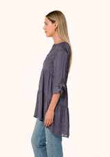 [Color: Purple Haze] A side facing image of a blonde model wearing a silky bohemian tunic top in a dusty purple diamond jacquard. With long rolled sleeves, a button tab sleeve closure, a v neckline, a tiered body, and a self covered button front. 