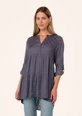 [Color: Purple Haze] A front facing image of a blonde model wearing a silky bohemian tunic top in a dusty purple diamond jacquard. With long rolled sleeves, a button tab sleeve closure, a v neckline, a tiered body, and a self covered button front. 