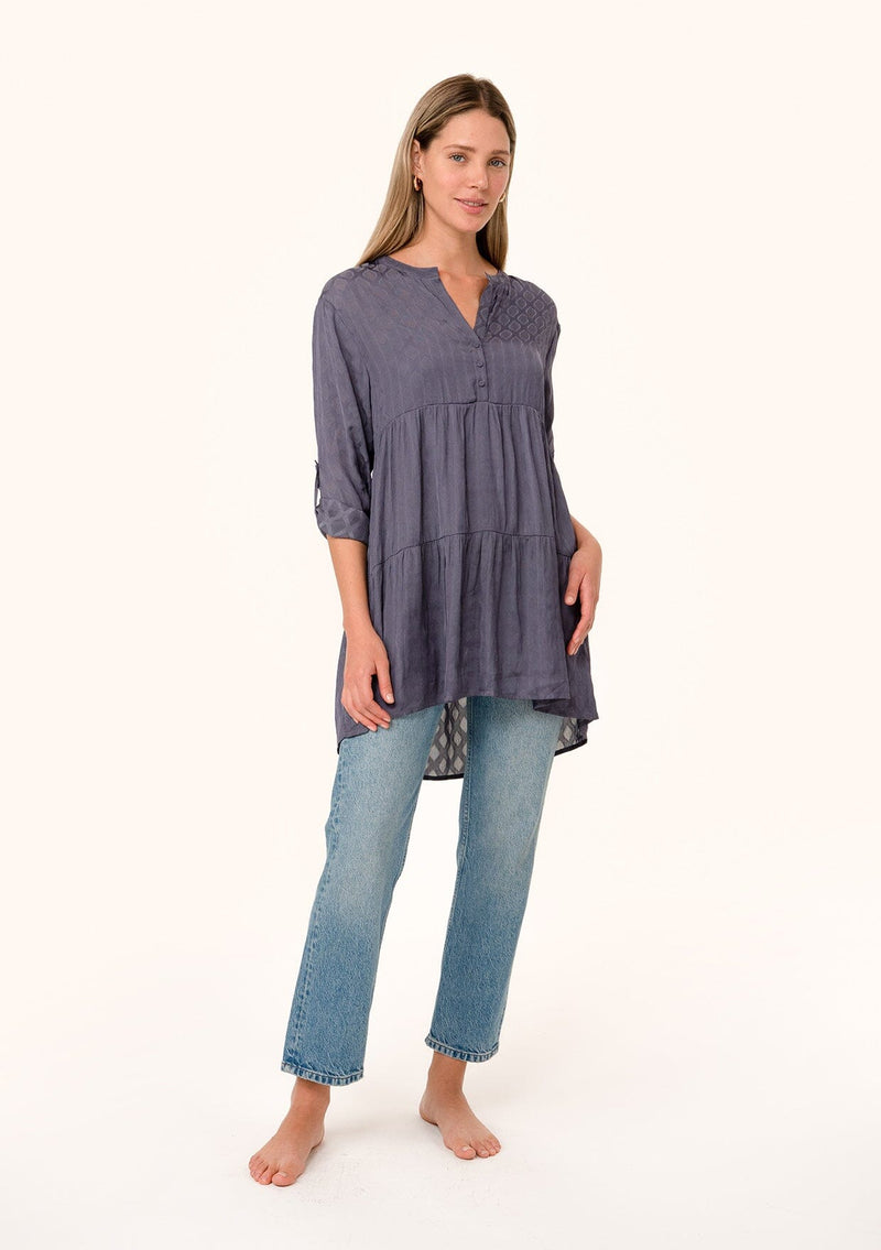[Color: Purple Haze] A full body front facing image of a blonde model wearing a silky bohemian tunic top in a dusty purple diamond jacquard. With long rolled sleeves, a button tab sleeve closure, a v neckline, a tiered body, and a self covered button front. 