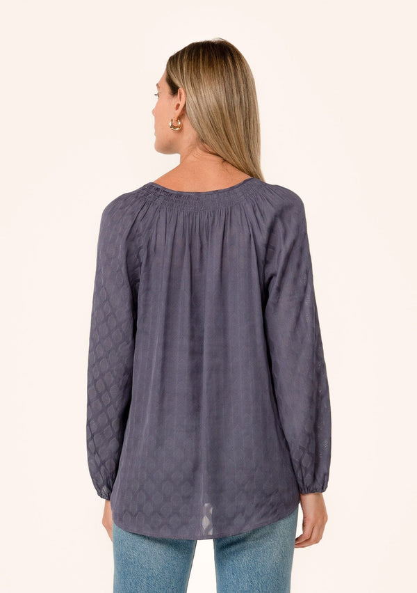 [Color: Purple Haze] A back facing image of a blonde model wearing a flowy fall bohemian blouse in a dusty purple diamond jacquard. With voluminous long raglan sleeves, a smocked neckline detail, a self covered button front, and a split v neckline with tassel ties. 