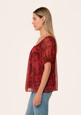 [Color: Wine/Charcoal] A side facing image of a blonde model wearing a fall chiffon blouse in a dark red floral and leaf print. With short puff sleeves, an elastic round neckline, and a relaxed, flowy fit.