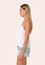 [Color: Chalk] A side facing image of a blonde model wearing a white bohemian camisole with adjustable spaghetti straps, a scoop neckline, a button up back, and lace detail.