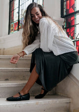 Woman sitting on steps in front of stained glass windows, wearing a white blouse and dark skirt.