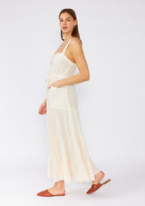 [Color: Vanilla] A side facing image of a brunette model wearing an off white linen blend sleeveless maxi dress. With a billowy tiered skirt, a large tortoise shell button front, an adjustable drawstring waist, and a cross back strap detail.