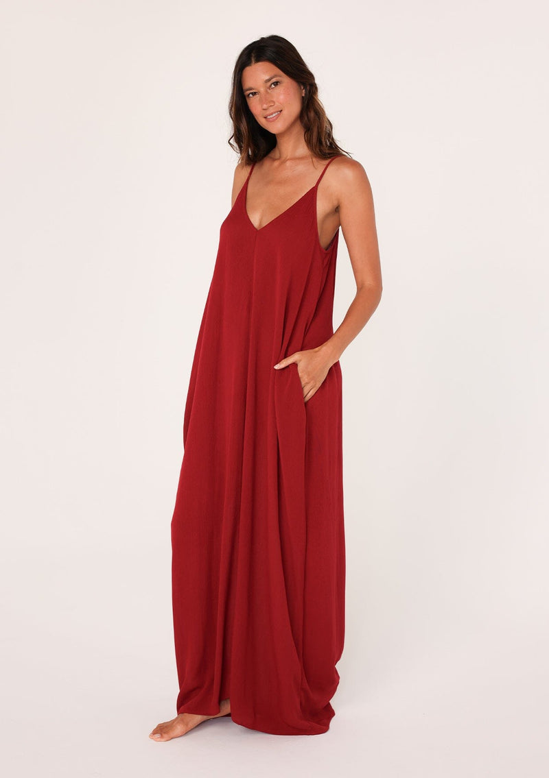 [Color: Brick Red] A front facing image of a brunette model wearing a red harem maxi dress. This billowy maxi tank top dress features a deep v neckline, adjustable spaghetti straps, and a cocoon fit.