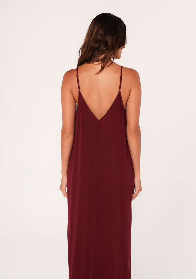 [Color: Merlot] A back facing image of a brunette model wearing a burgundy red harem maxi dress. This billowy maxi tank top dress features a deep v neckline, adjustable spaghetti straps, and a cocoon fit.