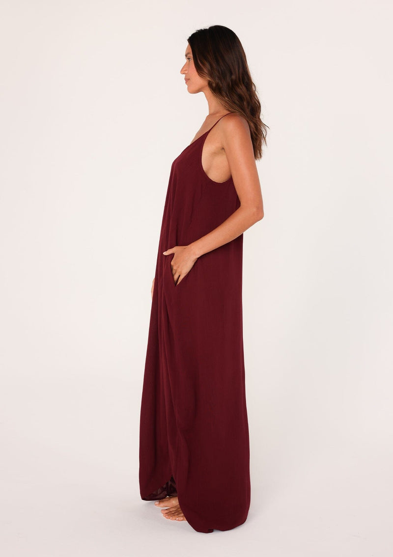 [Color: Merlot] A side facing image of a brunette model wearing a burgundy red harem maxi dress. This billowy maxi tank top dress features a deep v neckline, adjustable spaghetti straps, and a cocoon fit.