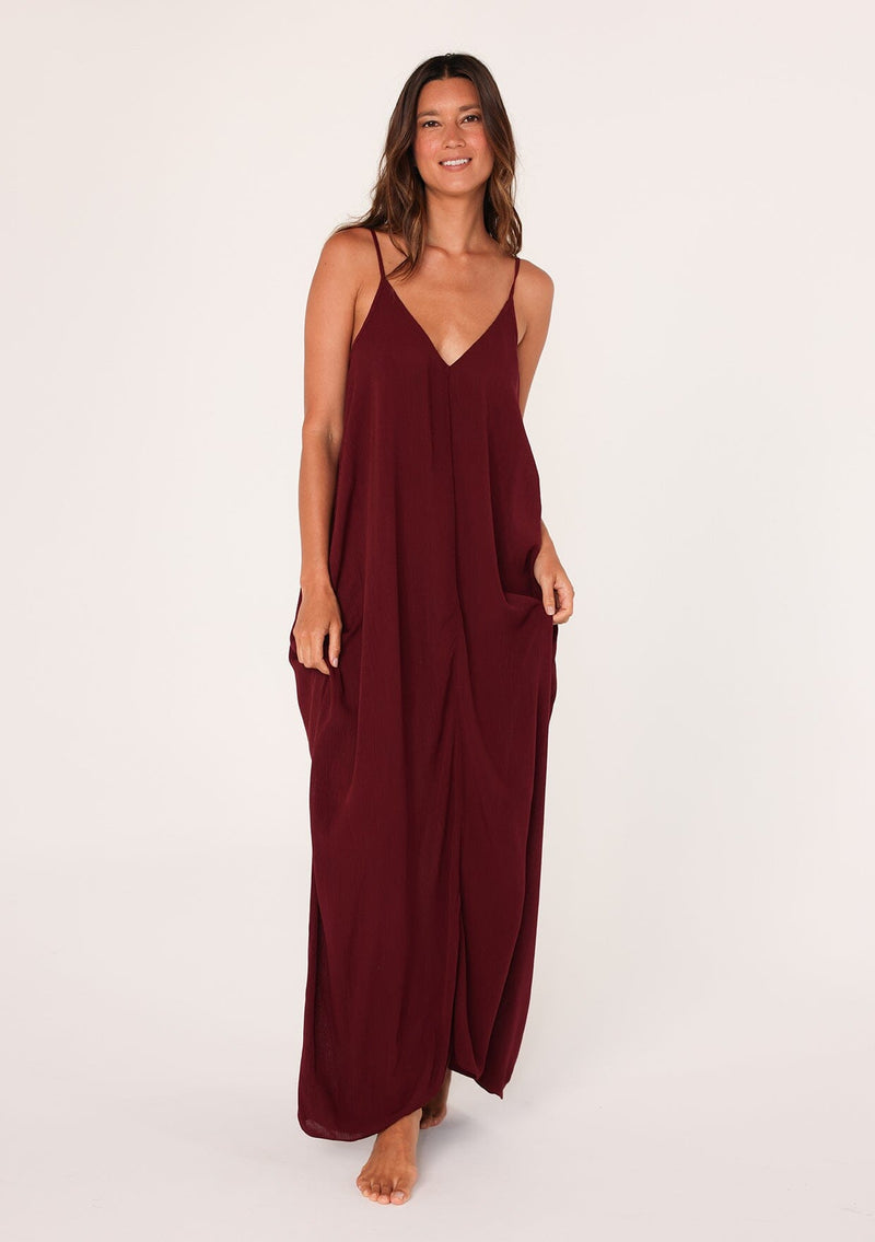 [Color: Merlot] A front facing image of a brunette model wearing a burgundy red harem maxi dress. This billowy maxi tank top dress features a deep v neckline, adjustable spaghetti straps, and a cocoon fit.