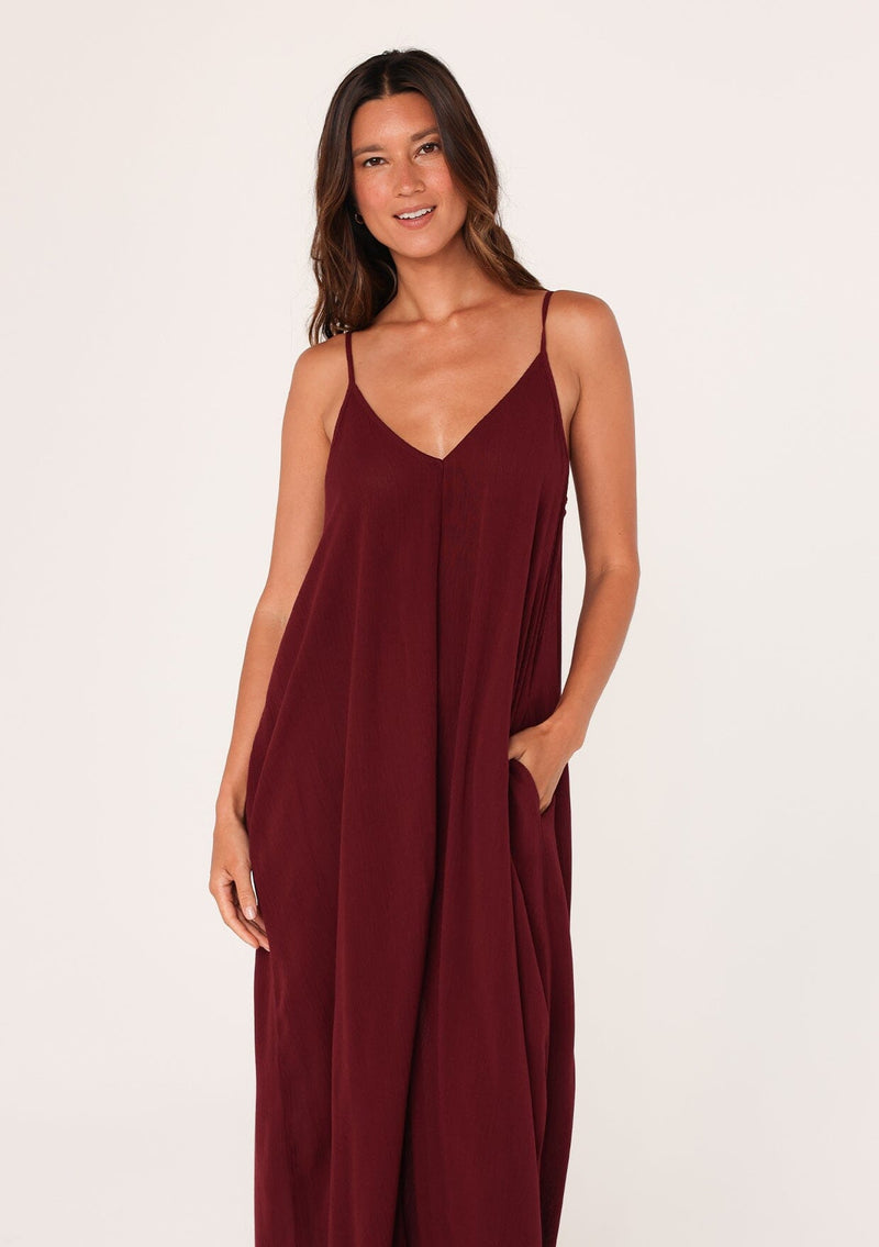 [Color: Merlot] A close up front facing image of a brunette model wearing a burgundy red harem maxi dress. This billowy maxi tank top dress features a deep v neckline, adjustable spaghetti straps, and a cocoon fit.