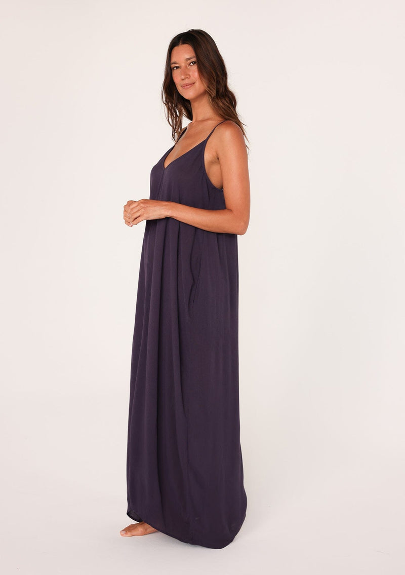 [Color: Dark Purple] A side facing image of a brunette model wearing a dark purple harem maxi dress. This billowy maxi tank top dress features a deep v neckline, adjustable spaghetti straps, and a cocoon fit.