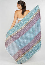 [Color: Blue/Green] A woman shown from the back wearing a white bikini top and holding up a large patterned beach towel. The fabric has horizontal stripes in light blue and lavender-purple with white fringe along the bottom edge.