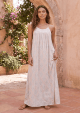 [Color: Nude Combo] A brunette model standing near a concrete wall and greenery wearing a bohemian maxi dress with a a halter neckline and a racerback with tassel tie detail. 