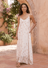 [Color: Blue Rose/Vanilla] A woman modeling a long, sleeveless  dress with thin straps and a delicate pink butterfly print set against a rustic stone archway and tiled patio.