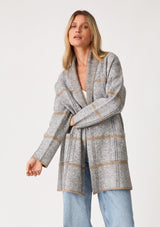 [Color: Grey/Taupe] A front facing image of a blonde model wearing a cozy fall cardigan in a grey windowpane plaid. With long sleeves, an open front, a shawl collar, and side pockets. 