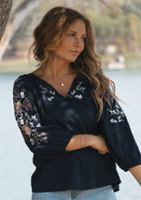 [Color: Eclipse/Coral] A front facing image of a blonde model by a lake wearing a dark blue embroidered blouse with 3/4 length sleeves,  a split v-neckline with tie details, and lattice trim accents. 