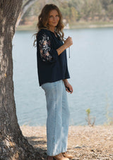 [Color: Eclipse/Coral] A side image of a blonde model by a lake wearing a dark blue embroidered blouse with 3/4 length sleeves,  a split v-neckline with tie details, and lattice trim accents. 