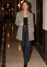 [Color: Cream/Black] A woman walking down a grand, warmly lit hotel hallway wearing a black and beige herringbone patterned blazer over a black top and dark blue jeans.