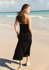 [Color: Black] A back facing image of a brunette model at the beach wearing a black knit halter dress with pointelle detail. Featuring a chic halter neckline, ruched bust with adjustable ties, and a front cutout. 