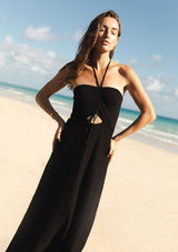 [Color: Black] A front facing image of a brunette model at the beach wearing a black knit halter dress with pointelle detail. Featuring a chic halter neckline, ruched bust with adjustable ties, and a front cutout. 
