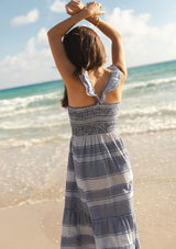 [Color: Navy/White] A back facing image of a brunette model at the beach wearing a blue and white checkered midi dress with stripes. Featuring chic ruffled elastic straps, a smocked bust, side pockets, and a tiered skirt. 