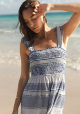 [Color: Navy/White] A detail image of a brunette model at the beach wearing a blue and white checkered midi dress with stripes. Featuring chic ruffled elastic straps, a smocked bust, side pockets, and a tiered skirt. 