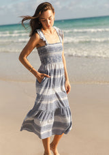 [Color: Navy/White] A side facing image of a brunette model at the beach wearing a blue and white checkered midi dress with stripes. Featuring chic ruffled elastic straps, a smocked bust, side pockets, and a tiered skirt. 