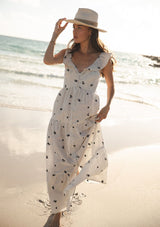 [Color: Natural/Grey] A front facing image of a brunette model at the beach wearing white and grey striped maxi dress with floral embroidery. Featuring flutter sleeves, a button front, a tiered skirt, and a smocked back. 