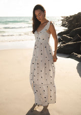 [Color: Natural/Grey] A side facing image of a brunette model at the beach wearing white and grey striped maxi dress with floral embroidery. Featuring flutter sleeves, a button front, a tiered skirt, and a smocked back. 