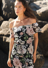 [Color: Black/Blush] A close up of a brunette model near rocks wearing a black and pink floral chiffon midi dress with an elastic off shoulder, braided waist belt, and tiered flowy skirt. 