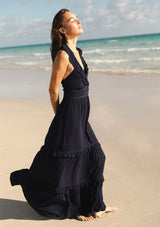 [Color: Navy] A side facing image of a brunette model at the beach wearing a flowy navy maxi dress with a halter neck, v-neckline, ruffle details, and a tiered skirt. 
