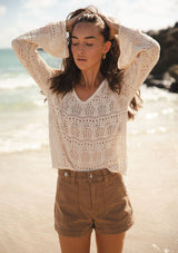 [Color: Natural] A front facing image of a brunette model at the beach wearing a beige crochet knit top with long bell sleeves and a v-neckline. A breezy beach top for spring and summer. 