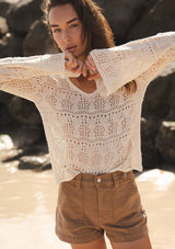 [Color: Natural] A front facing image of a brunette model at the beach wearing a beige crochet knit top with long bell sleeves and a v-neckline. A breezy beach top for spring and summer. 