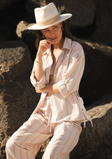 [Color: Sand/White] A front facing image of a brunette model at the beach sitting on rocks wearing a beige and white striped shirt with a collared neckline, button front, roll tab sleeves, and a playful side tie detail. 