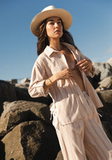 [Color: Sand/White] A front facing image of a brunette model at the beach by rocks wearing a beige and white striped shirt with a collared neckline, button front, roll tab sleeves, and a playful side tie detail. 