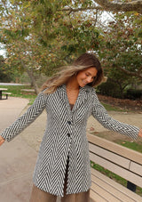 [Color: Cream/Black] A front facing image of a brunette model wearing a classic women's coat in a cream and black chevron stripe. With long sleeves, a notched collar, a button front, and side pockets. 