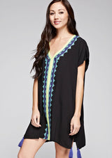 [Color: Black] A side facing image of a brunette model wearing a black embroidered tunic with embroidery and a tassel accent at the hem.