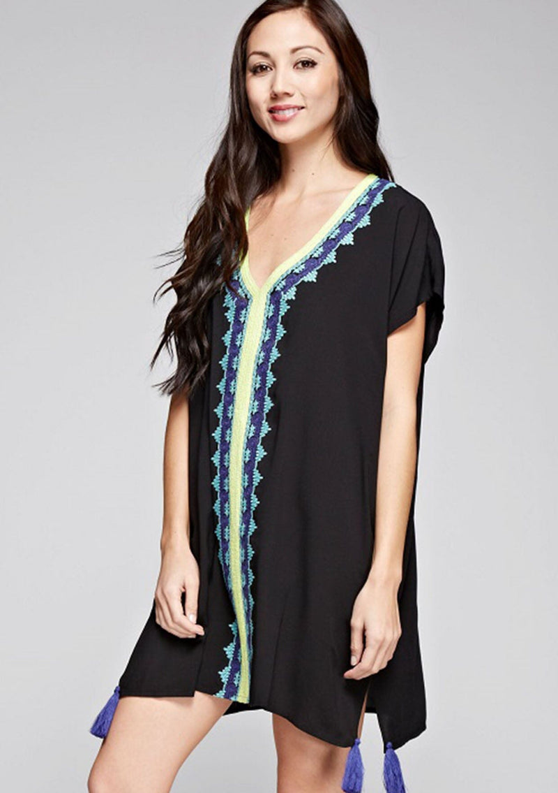 [Color: Black] A side facing image of a brunette model wearing a black embroidered tunic with embroidery and a tassel accent at the hem.
