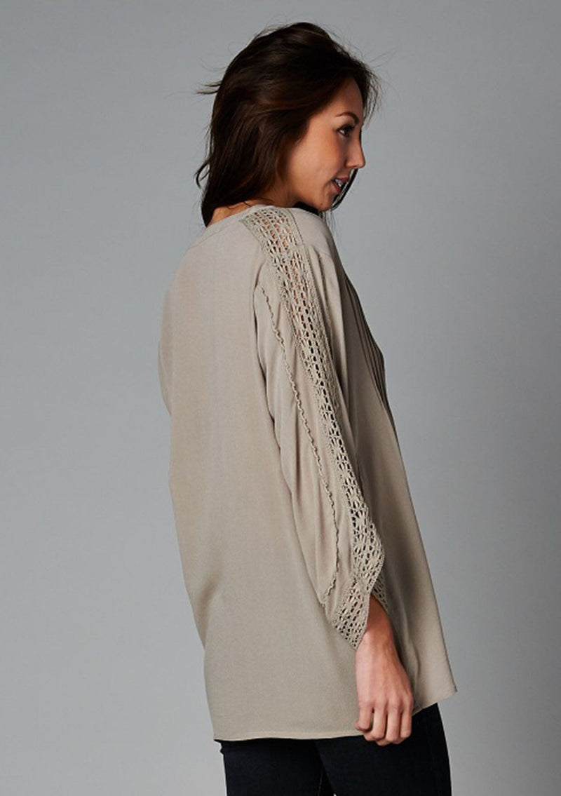 [Color: Taupe] A side facing image of a brunette model wearing a bohemian v-neck blouse with lace crochet detail at the sleeve.